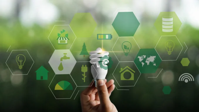 A hand holds a low-energy light bulb in front of a green hexagonal grid of sustainability icons covering renewable energy, water, recycling and buildings.