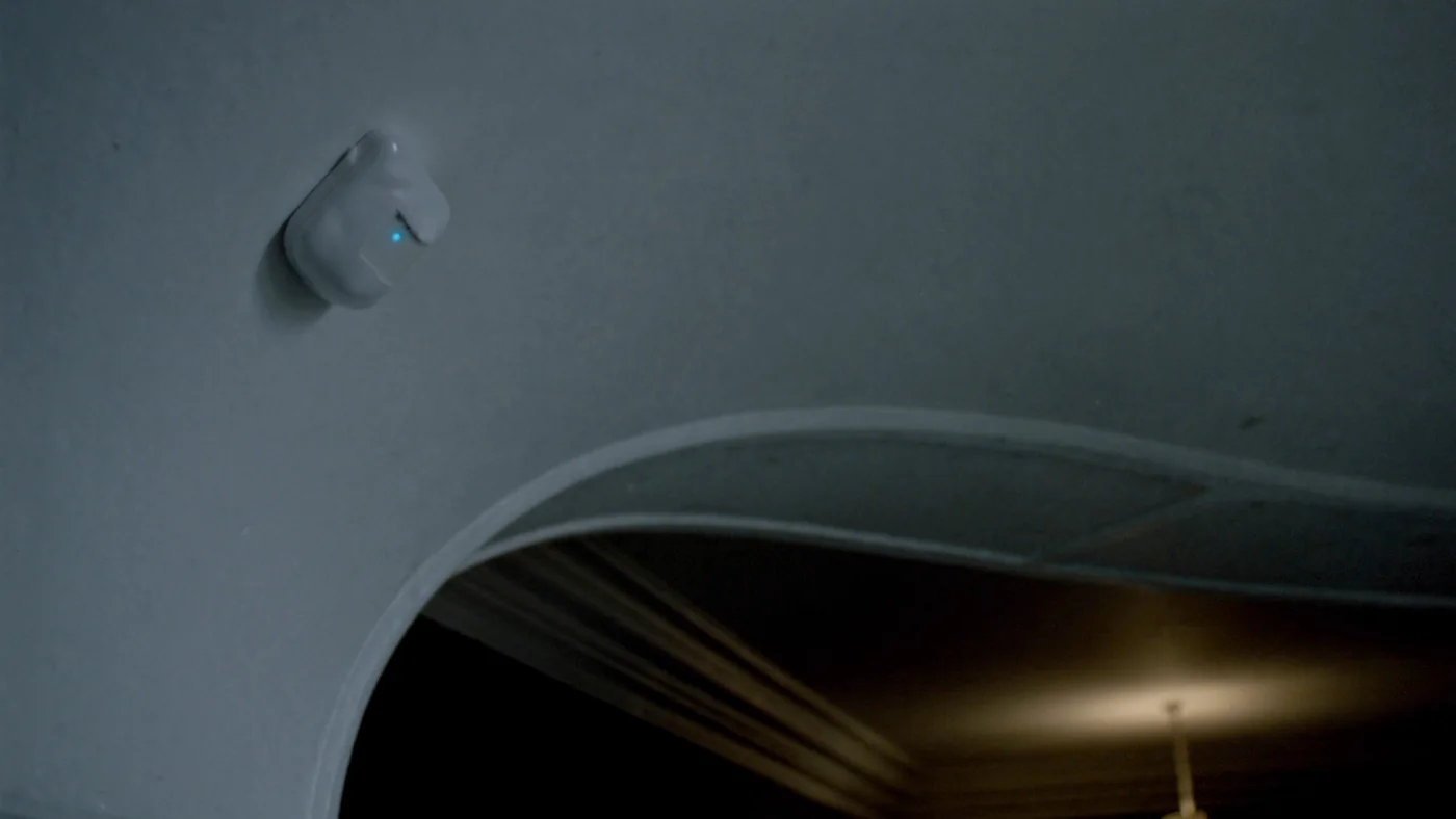 A small white wireless sensor mounted high on a corridor wall, status light glowing faintly